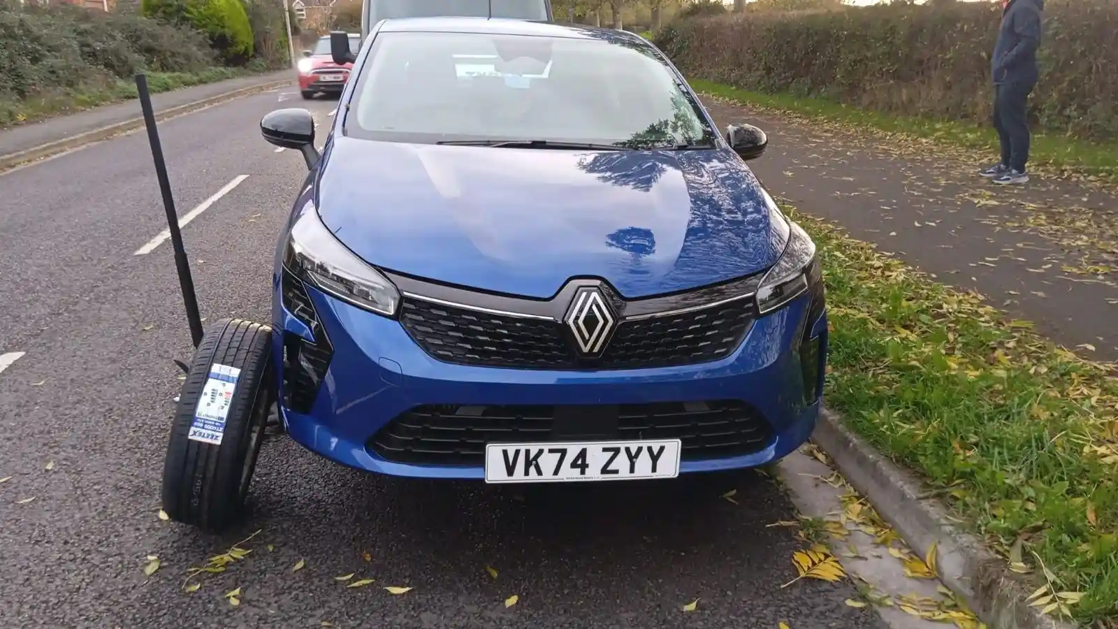 A blue Renault Clio is parked on the side of a road with a detached tire nearby
